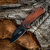 Nóż składany Fox Outdoors Compact – drewno egzotyczne - rozłożony, obok drewna w terenie, ostrzem skierowanym w dół